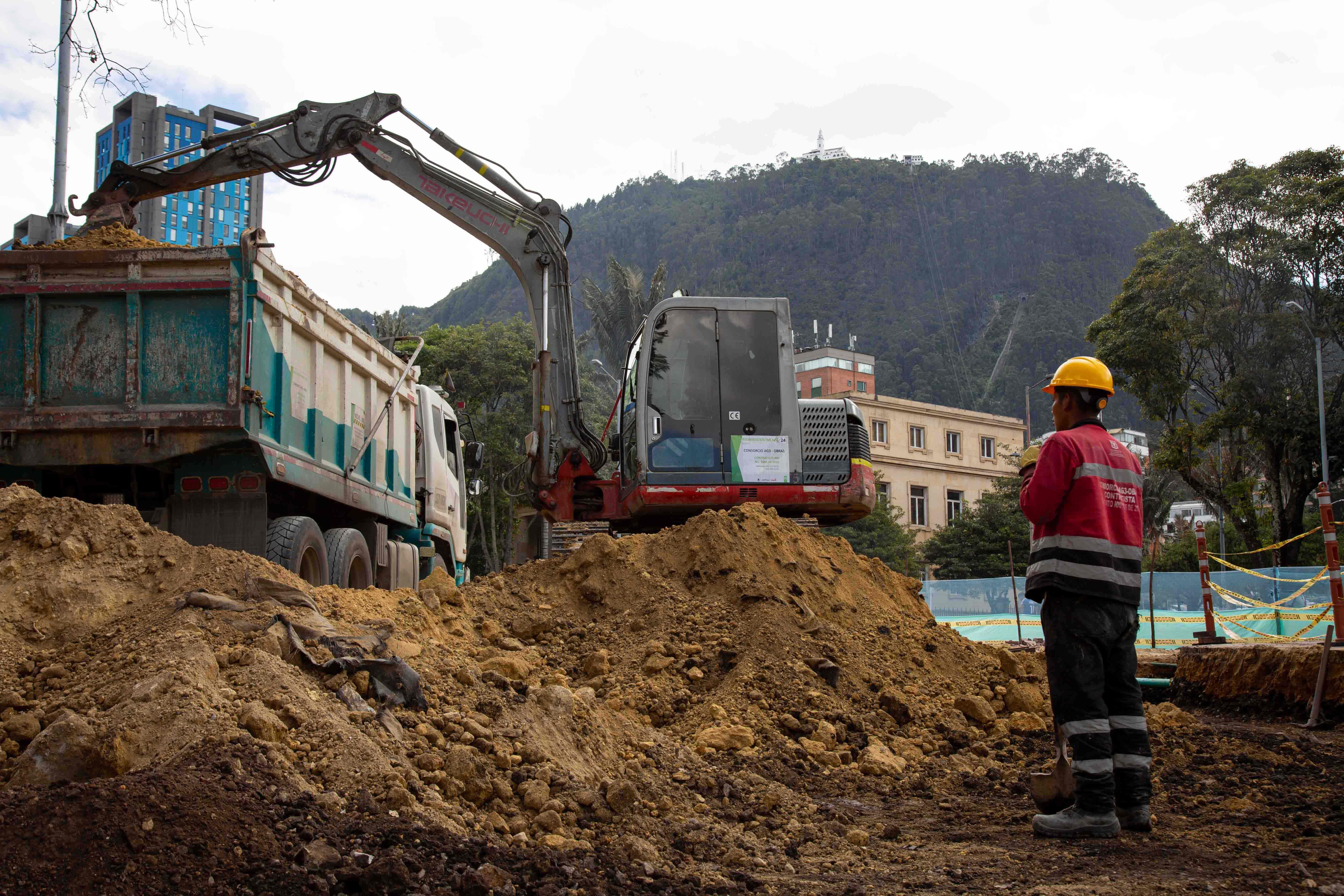Un empleado de obra está parado al lado de una montaña de tierra y una máquina excavadora. Al fonde está Monserrate