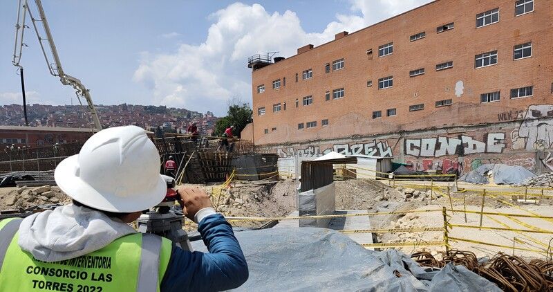 Un topógrafo realiza la medición mientras al fondo se ve un trabajador sobre un puente en construcción