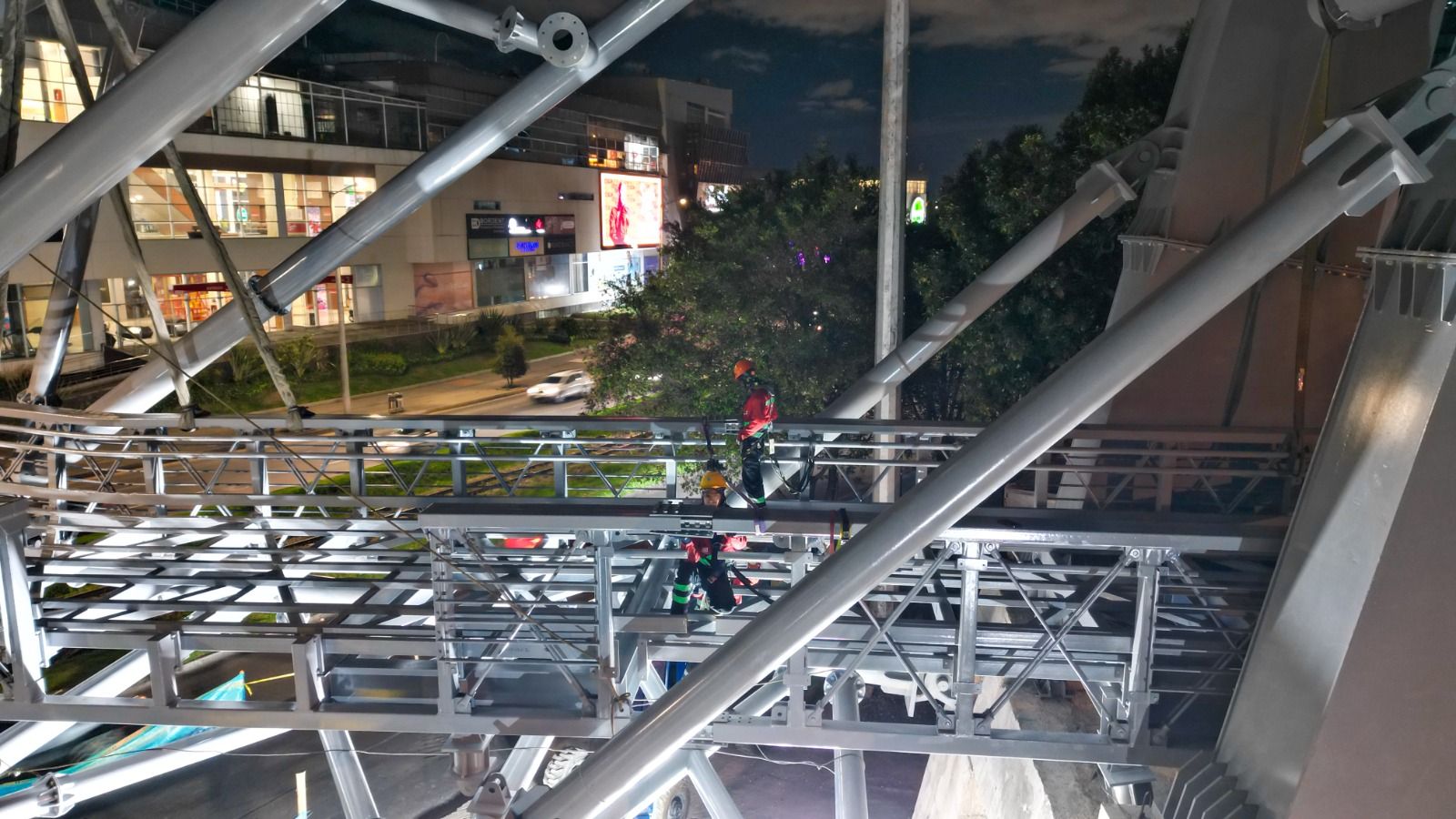 Trabajadores con arnés y cascos de protección realizan labores de soldadura y ajuste sobre la pasarela metálica de noche.